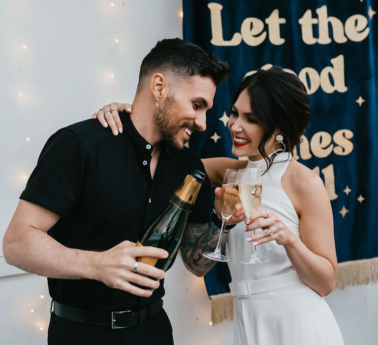 Bride in a halter neck wedding dress with belt detail and front split sipping champagne with her groom in a black shirt 