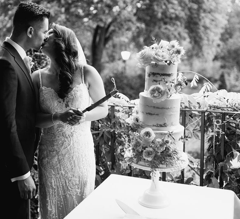 Bride in a lace fitted wedding dress kissing her groom after they've cut their semi-naked wedding cake 