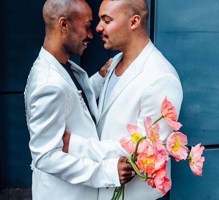 two grooms in white satin jackets embracing whilst holding a red anemone bouquet 