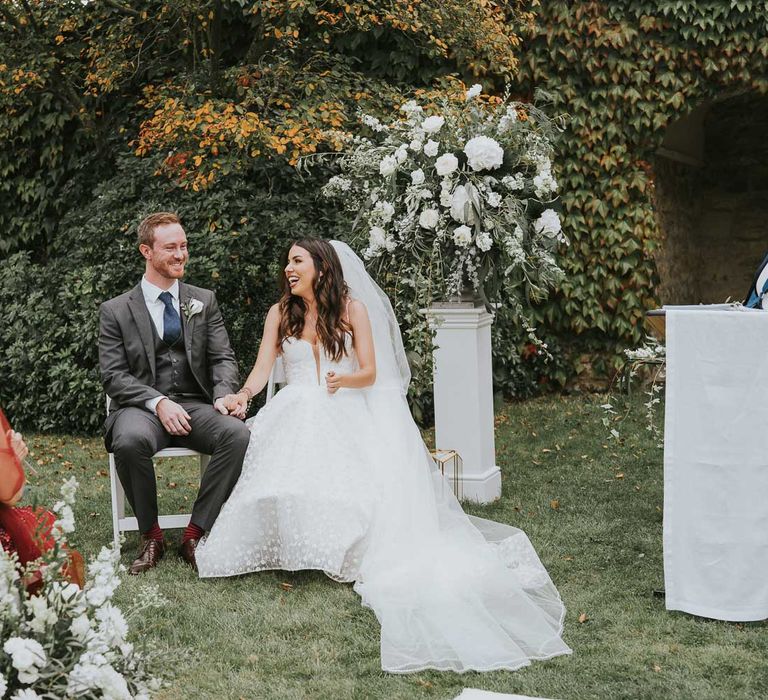 Bride in Pronovias wedding dress, Jimmy Choo wedding shoes and veil sits on white wooden chair during outdoor wedding ceremony with groom in grey three piece wedding suit, blue tie and white rose buttonhole