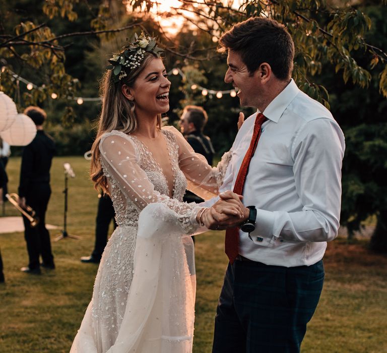 Bride in long sleeve v neck wedding dress and flower crown dances during golden hour with groom in white shirt a red tie at tipi wedding reception