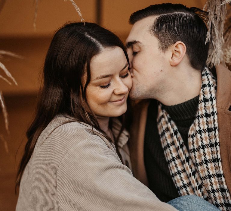 Groom-to-be in a rust coloured coat and check scarf kissing his bride-to-be's cheek at their boho wedding proposal 