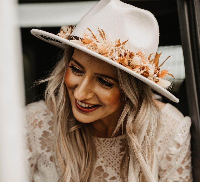 Smiling bride in white bridal fedora with orange dried flower details sits in car wearing lace boho bridal top