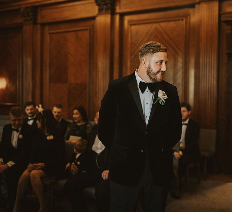 Groom in a tuxedo with bow tie and pink buttonhole flower waiting at the altar of his Old Marylebone Town Hall wedding 