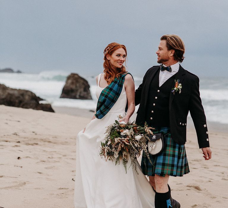 Bride in a princess wedding dress with tartan sash and groom in a kilt walking along the beach at Big Sur, California 