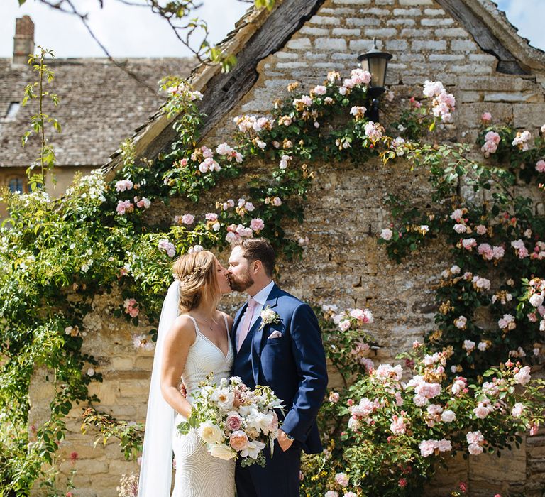 Summer wedding bride and groom portrait by a climbing rose bush with groom in a navy suit and bride and beaded wedding dress