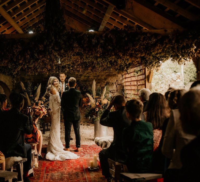 Bride in floral applique wedding dress with train stands with groom in dark suit during wedding ceremony with rustic decor