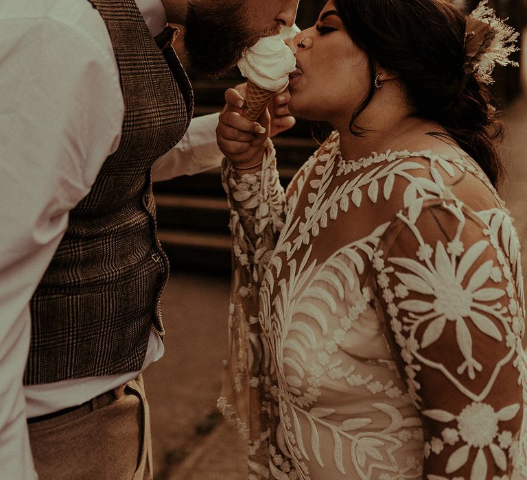 Groom in a tweed waistcoat and bride in a long sleeve lace wedding dress eating ice-cream together 