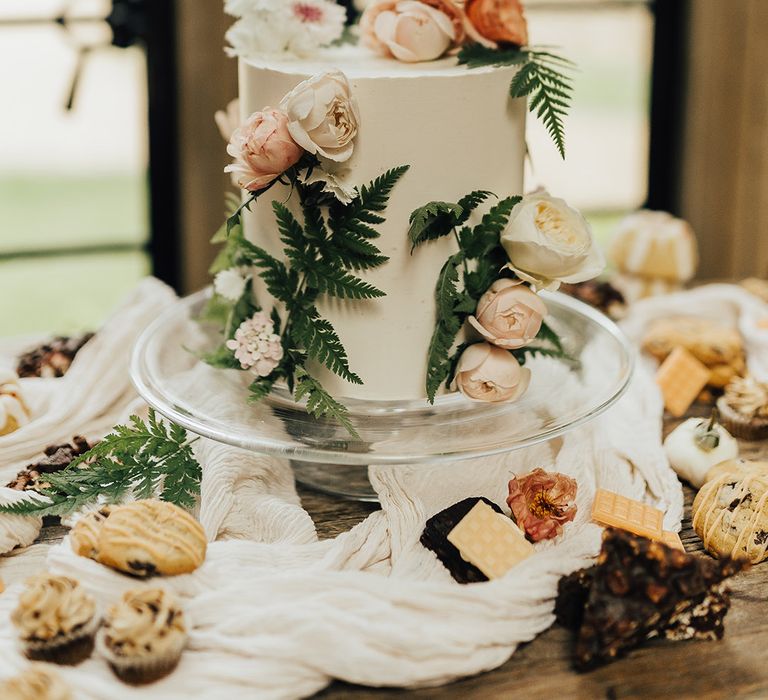 Single tier buttercream wedding cake on a glass cake stand decorated with flowers and fern leafs