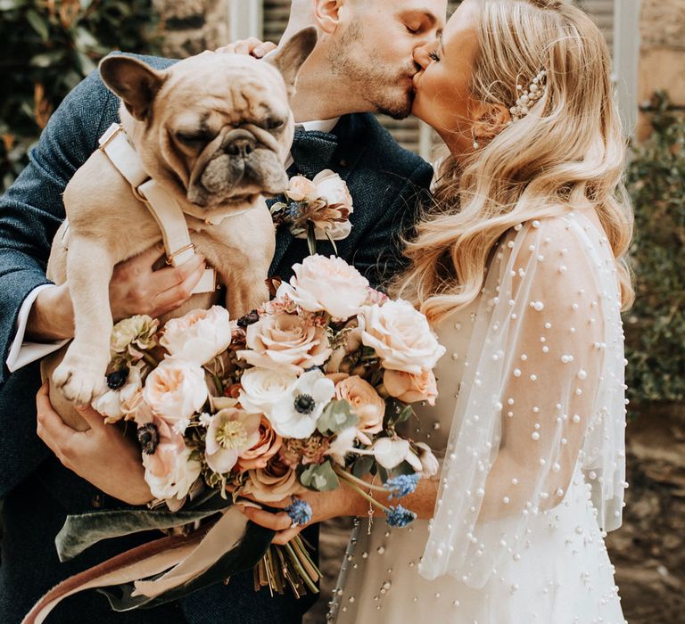 Bride in pearl wedding dress with sleeves kisses groom in navy tweed wedding suit holding French bulldog 