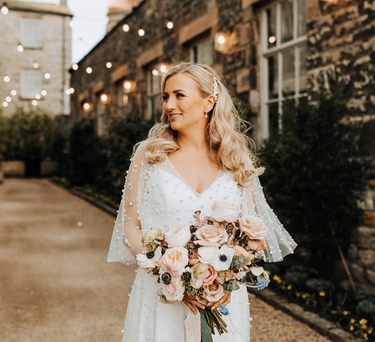 Bride with white short sleeve wedding dress with pearls stands holding mixed pastel wedding bouquet with roses and anemones