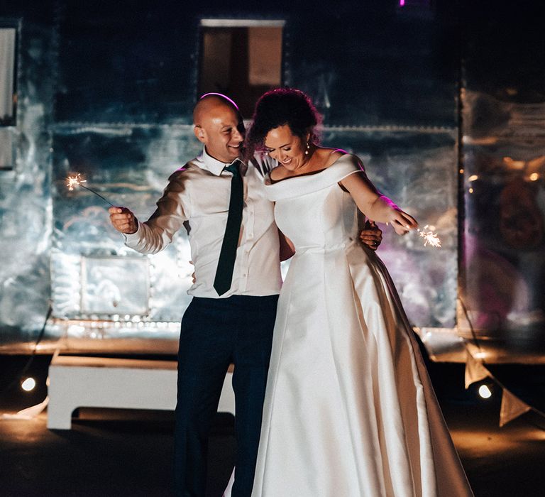 Bride and groom hold sparklers at their wedding reception under large neon pink sign reading 'photos' 