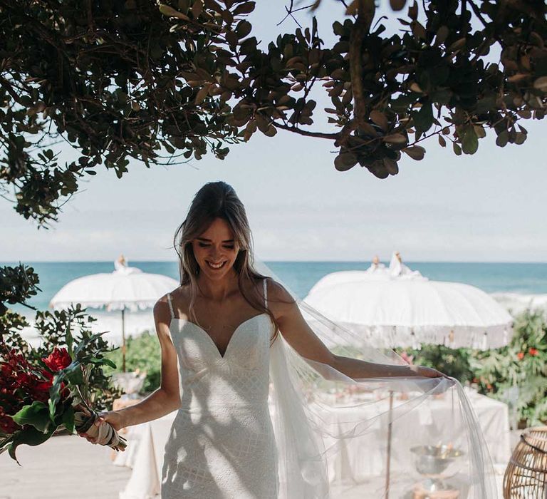 Bride in lace v-neck mermaid wedding dress with veil and red bouquet for beach ceremony