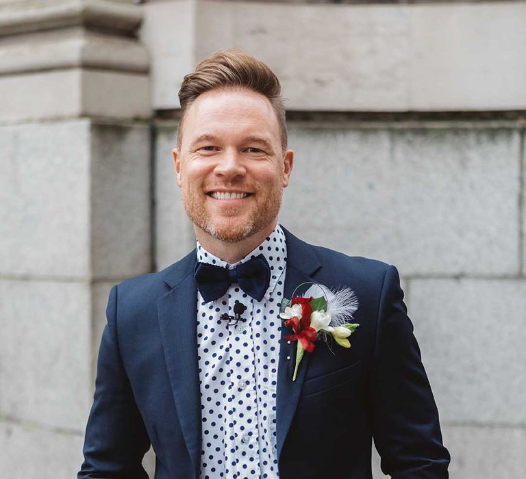 Groom wears deep blue suit with matching polka dot shirt and bow tie with floral buttonhole 