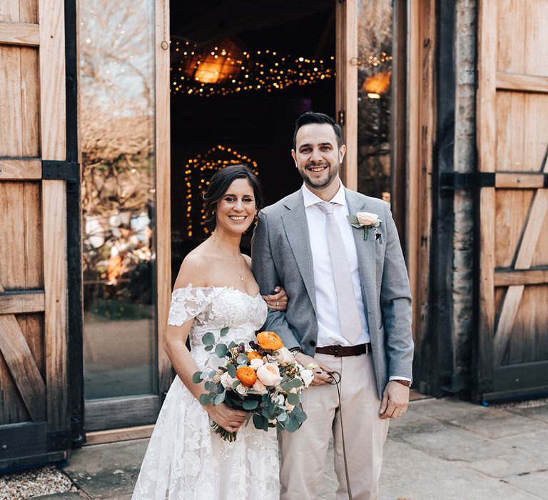 Bride and groom smile at the camera as the bride holds onto leash of their pet dog wearing love heart bow tie 