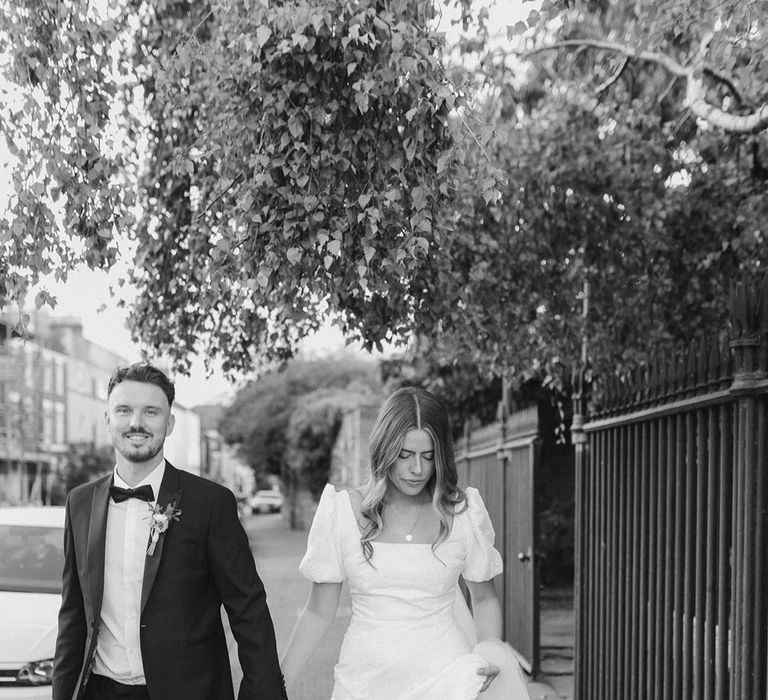 Groom in black tie intertwines his fingers with bride wearing Loeffler Randall white bow shoes