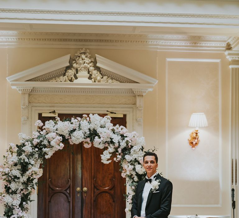 Pink and white wedding flower arch with groom in black tie waiting for the bride 