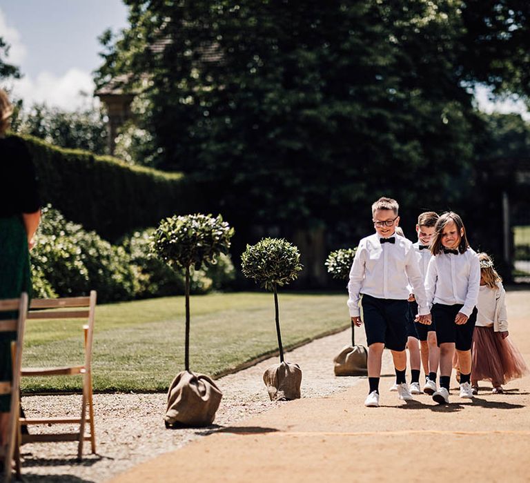 Page boys in black shorts, white shirt and black bow ties walks together followed by two flower girls in pink dresses and white flower crowns