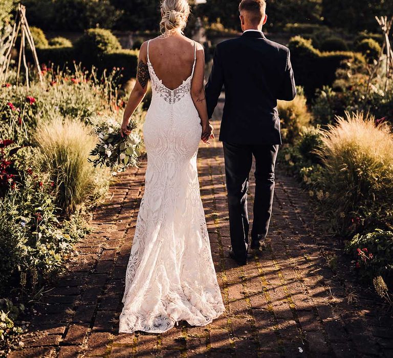 Bride in scoop back lace wedding dress with button details walks in the gardens holding hands with the groom in a blue tuxedo 