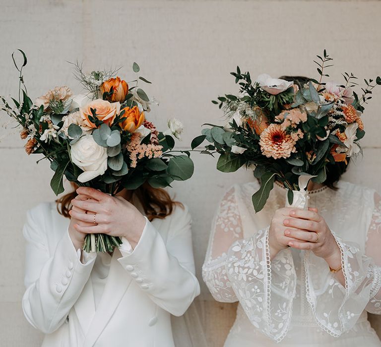 Brides hold up floral bouquets complete with pops of orange and white florals with green foliage