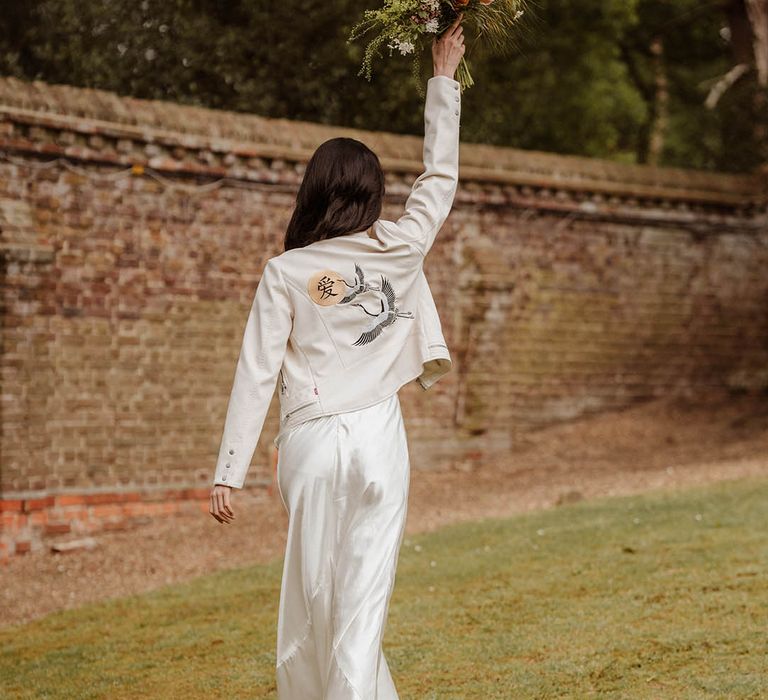 Bride in a satin slip wedding dress and white jacket with ancient Japanese motif holding her bouquet in the air at Gorse Hill