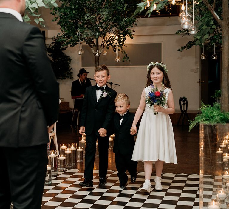 Page boys in black tie and flower girl in white dress with a white flower crown walk down the aisle together 