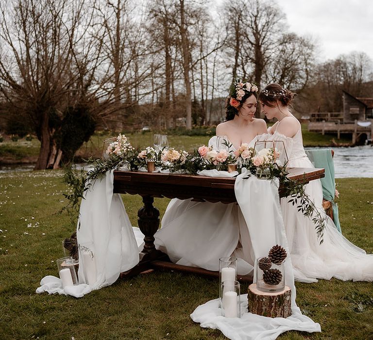 Two brides sitting at their outdoor sweetheart table decorated with drapes, pink flowers and lanterns in an ethereal wedding dress with tulle skirt and a strapless princess wedding dress with puff sleeves