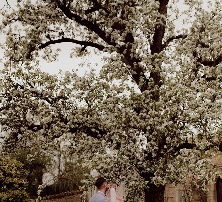 Bride wearing floor-length veil and Jane Hill wedding dress holds her groom beneath white blossom tree outdoors