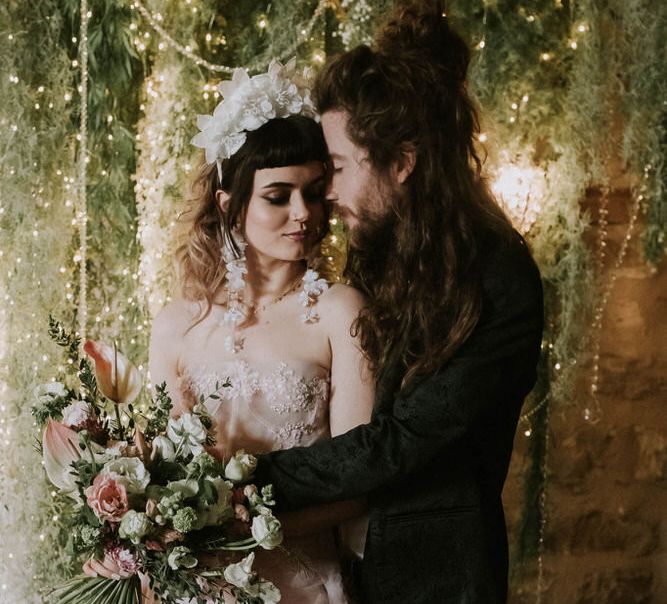 Groom in a black suit embracing his bride in a pink wedding dress as she holds a spring flower bouquet in front of a rustic twig, foliage and fairy light backdrop 