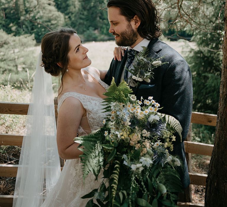 Bride holds oversized floral bouquet filled with foliage and daisies whilst she embraces her groom wearing three piece suit