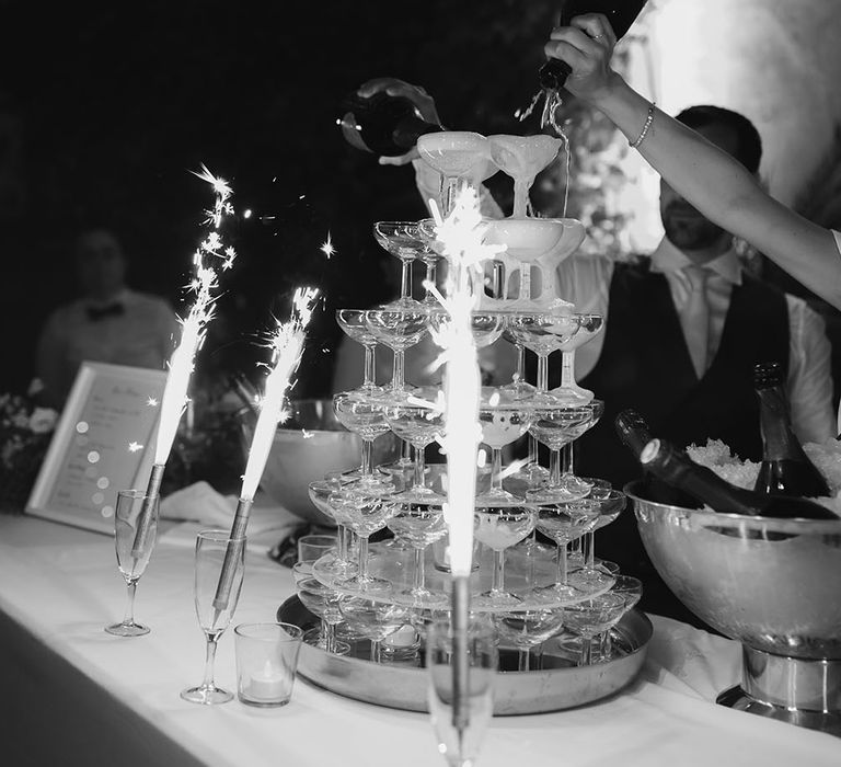 Bride & groom pour champagne into champagne tower in front of sparklers in glasses 