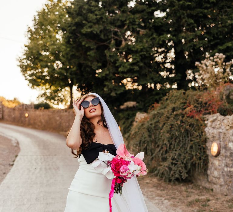 Bride wears white mini dress with black bow to front and cathedral veil whilst holding bright pink floral bouquet  