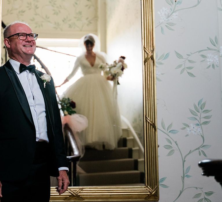 Bride wearing princess tulle wedding dress walks down staircase as her father wearing black tie awaits her at the bottom 