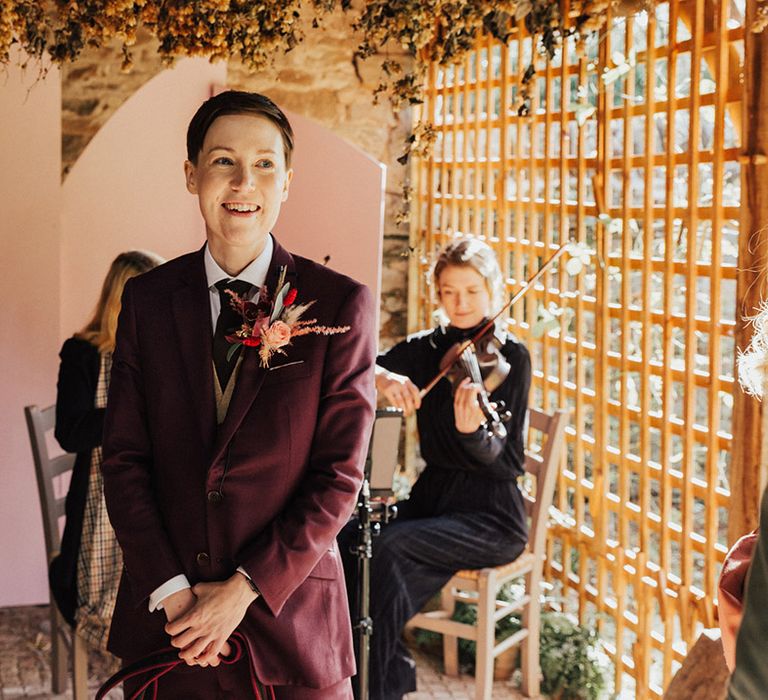 Ceri wears a burgundy suit with a dried flower buttonhole with fresh flowers 