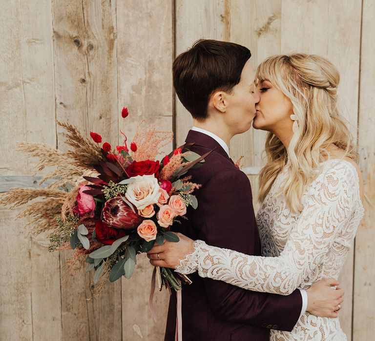 Bride in a long sleeve lace wedding dress kissing her partner who wears a burgundy suit 