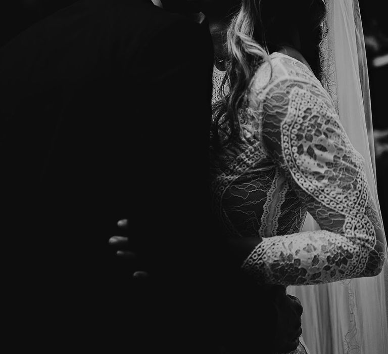 Groom in black tie leans in and kisses the bride on the cheek who wears a long sleeve boho lace wedding dress 