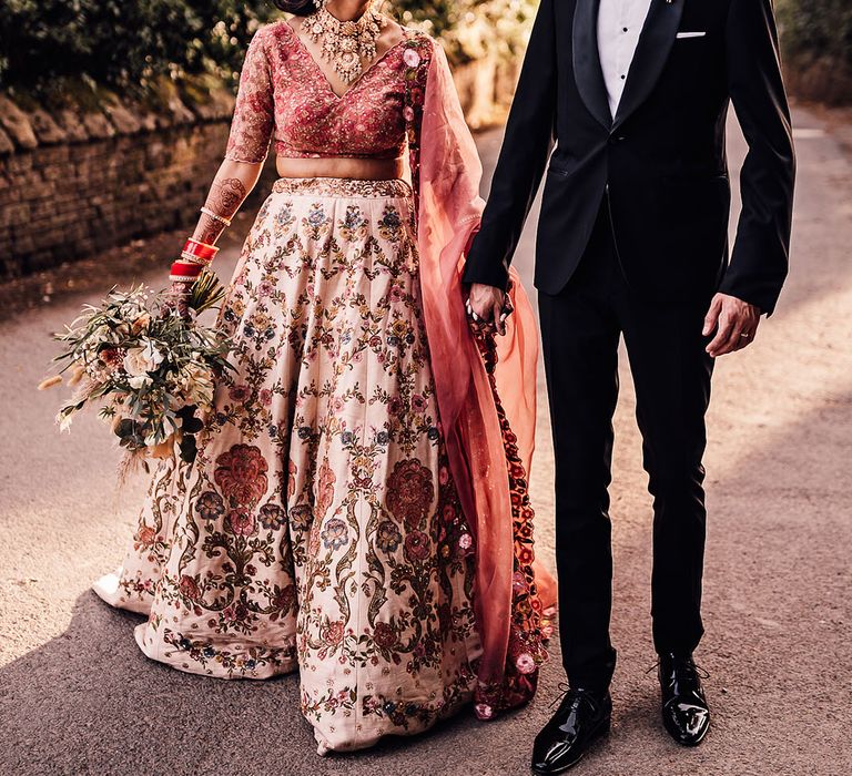 Bride wearing traditional pink lehenga with floral inspired design walks with her groom wearing black tie 