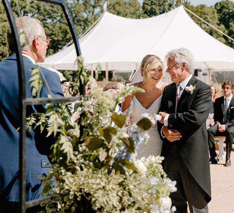 Bride stands beside her father who wears traditional morning suit during outdoor wedding in the summer