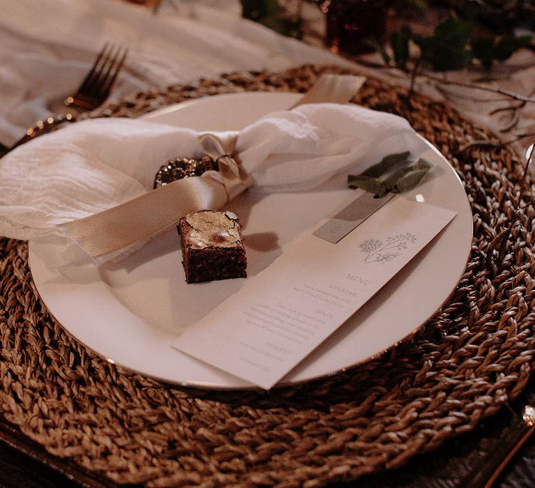 White napkin tied with pink ribbon, mini brownie favour with white stationery on a wicker placemat 