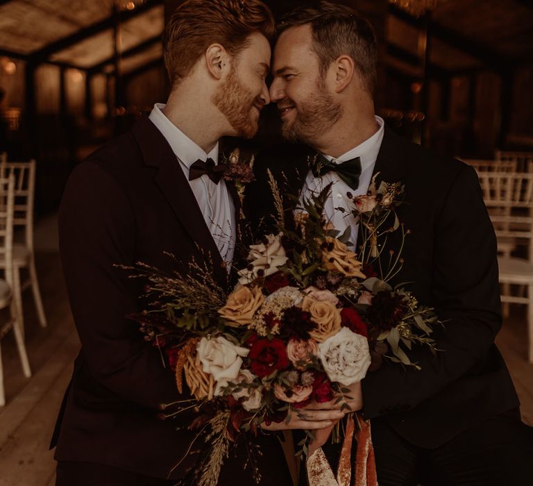 Grooms in black tie rest their foreheads against each other holding their stunning autumnal wedding bouquet 