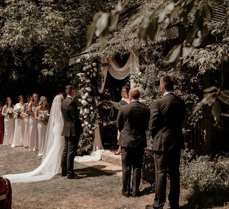 Bride and groom stand at the altar for the outdoor ceremony at Pennard House with blush pink flowers and bridesmaids in gold dresses