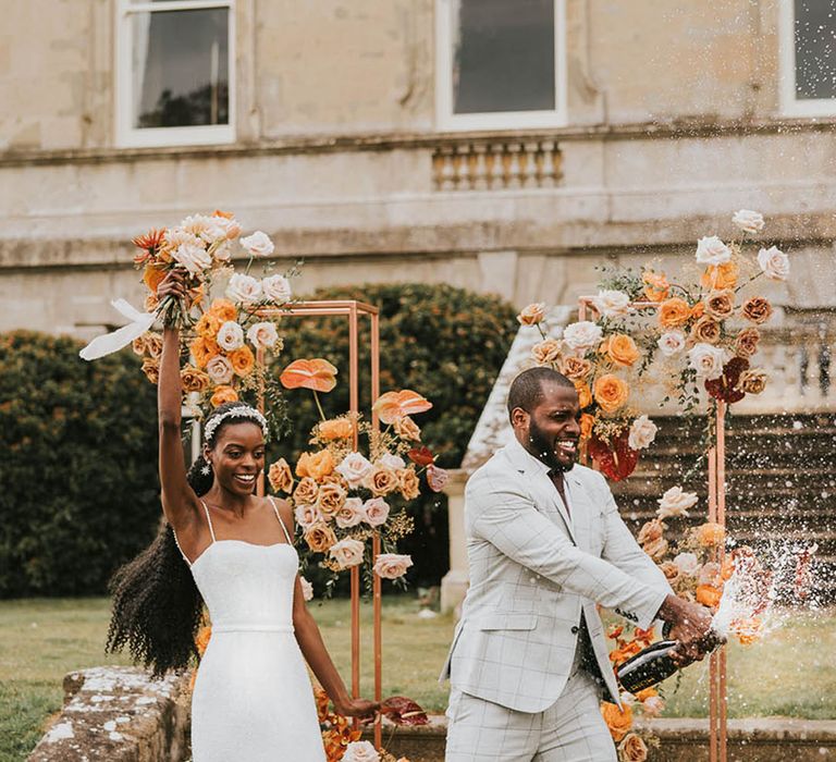 Black bride in a dress with pearl straps and the groom in a checkered suit popping a champagne bottle with orange and white roses 