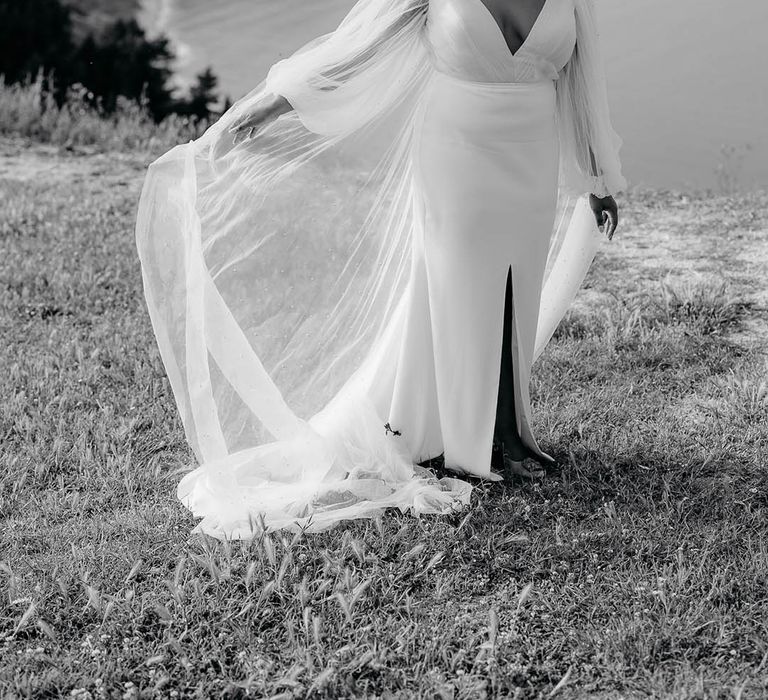 Bride shows of veil, sheer-sleeved wedding dress and something blue wedding shoes in front of ocean in Italy