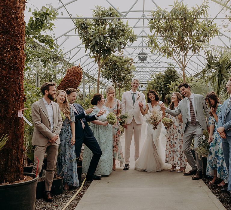 Posed photo of the wedding party in greenhouse with members pointing at each other and laughing