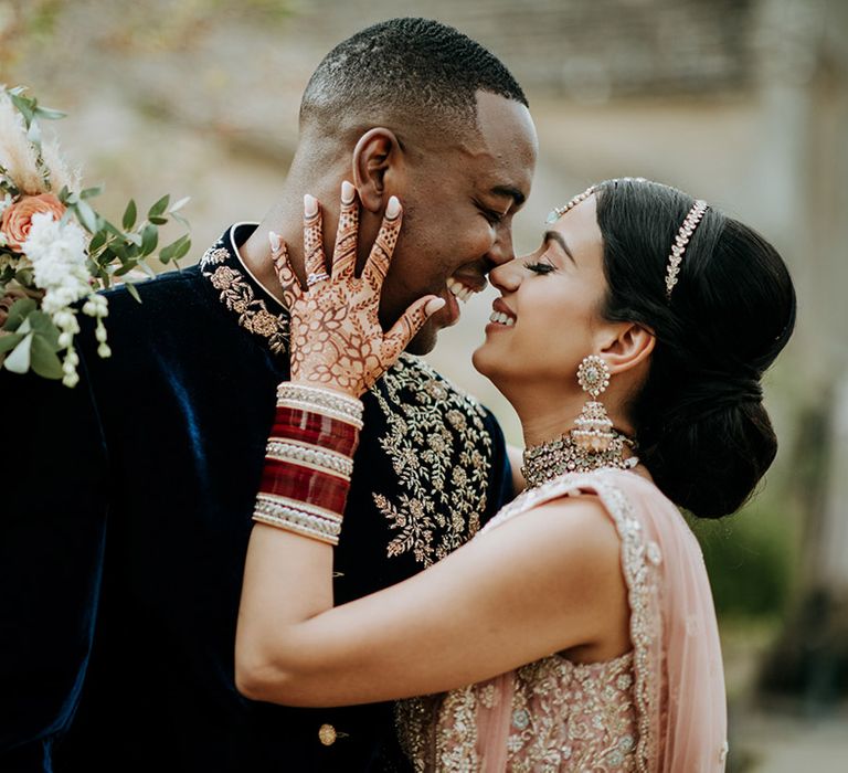 Panjabi Bride and Jamaican Groom pose at multicultural wedding. The bride wears a golden bridal Lehnga and carries a champagne bridal bouquet and the groom wears a gold embellished Indian wedding outfit