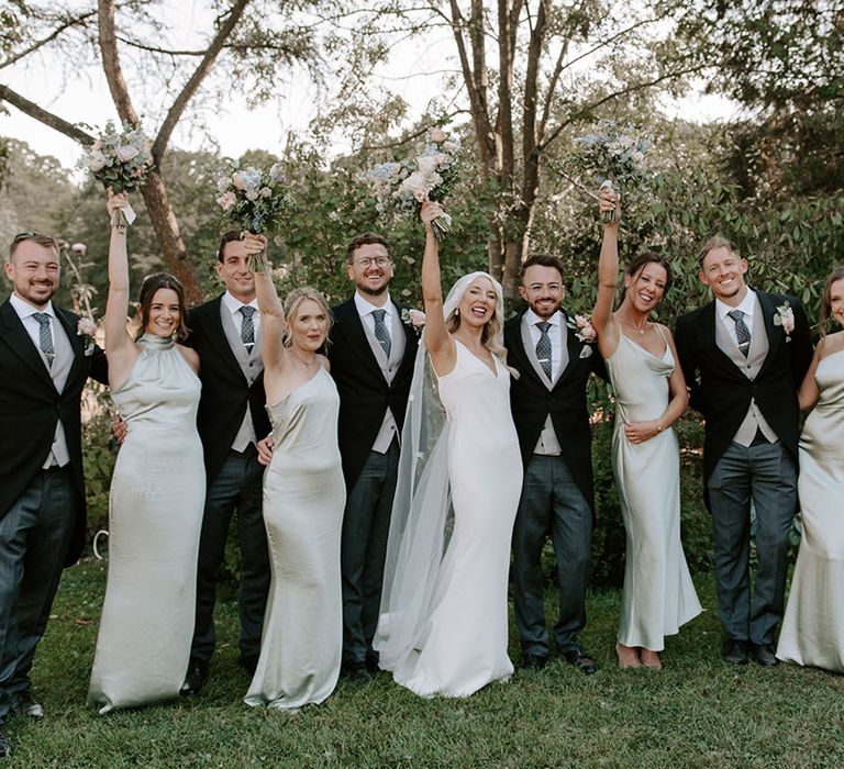 Bride and groom pose with their wedding party with bridesmaids in sage green dresses and groomsmen in three piece morning suits 