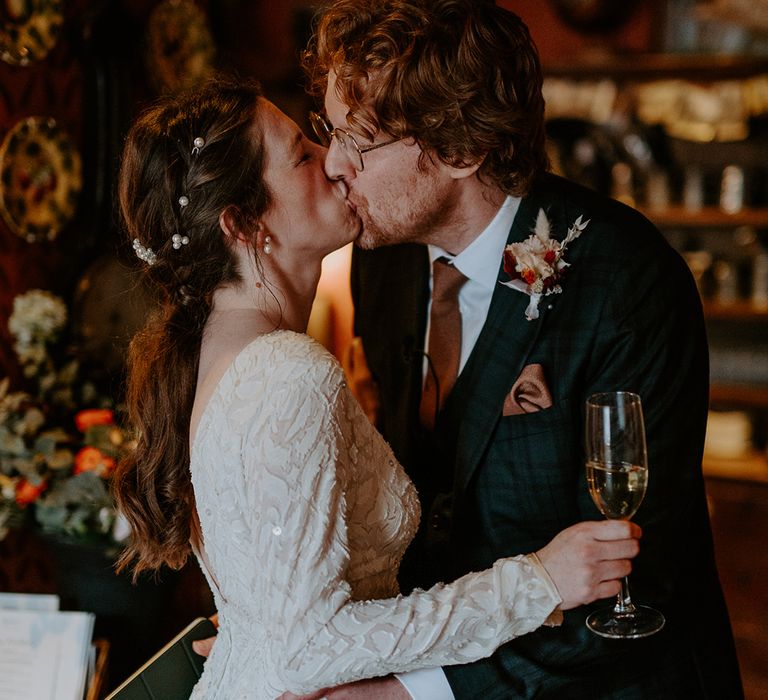 Bride in long sleeve lace vintage wedding dress, pearl earrings and pearl hair accessories kissing groom in dark suit with orange tie and autumnal boutonniere 