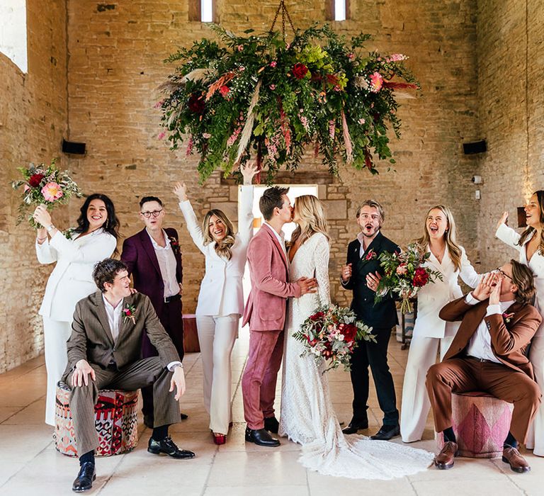 Bride and groom kissing standing by bridesmaids in matching white bridesmaid jumpsuits with white blazers and groomspeople in mismatched groom suits underneath suspended foliage, rose, pampas grass and eucalyptus decor