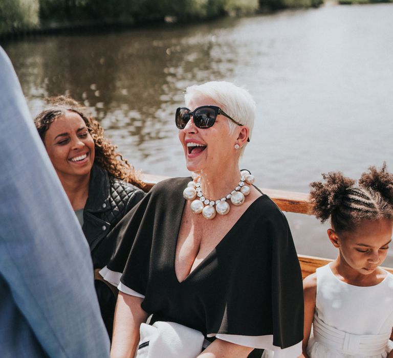 Wedding guest in statement necklace and sunglasses sits beside flower girl during Thames boat ride 