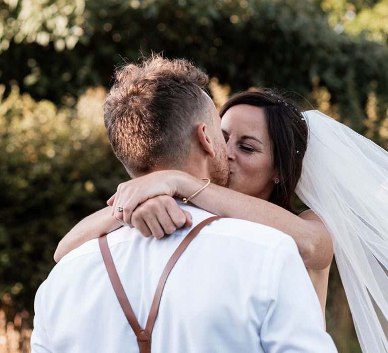 Bride in floor-length veil kisses her groom outdoors during couples portraits after rustic barn wedding 